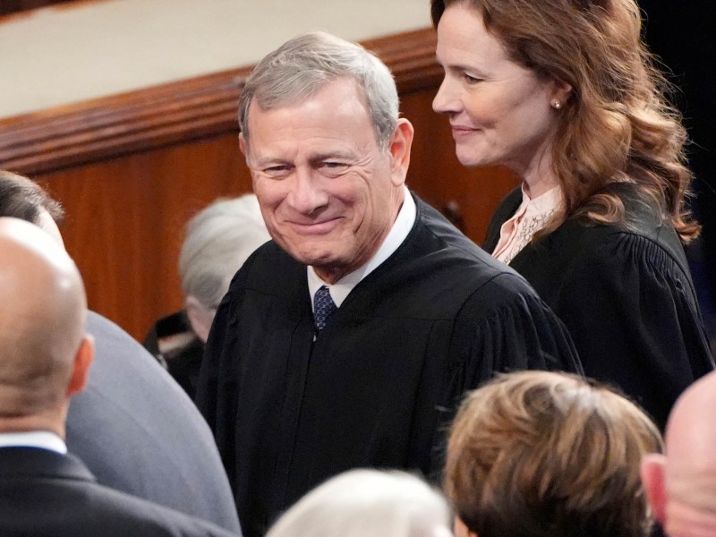 A closeup of Chief Justice of the Supreme Court John Roberts, in black robes, walking into a room of people