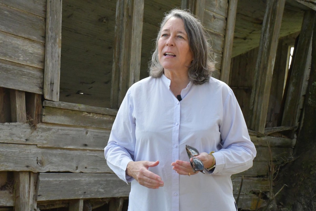 A woman stands in the center of the frame with a weathered structure behind her, looking to her right and gesturing with her hands as she speaks