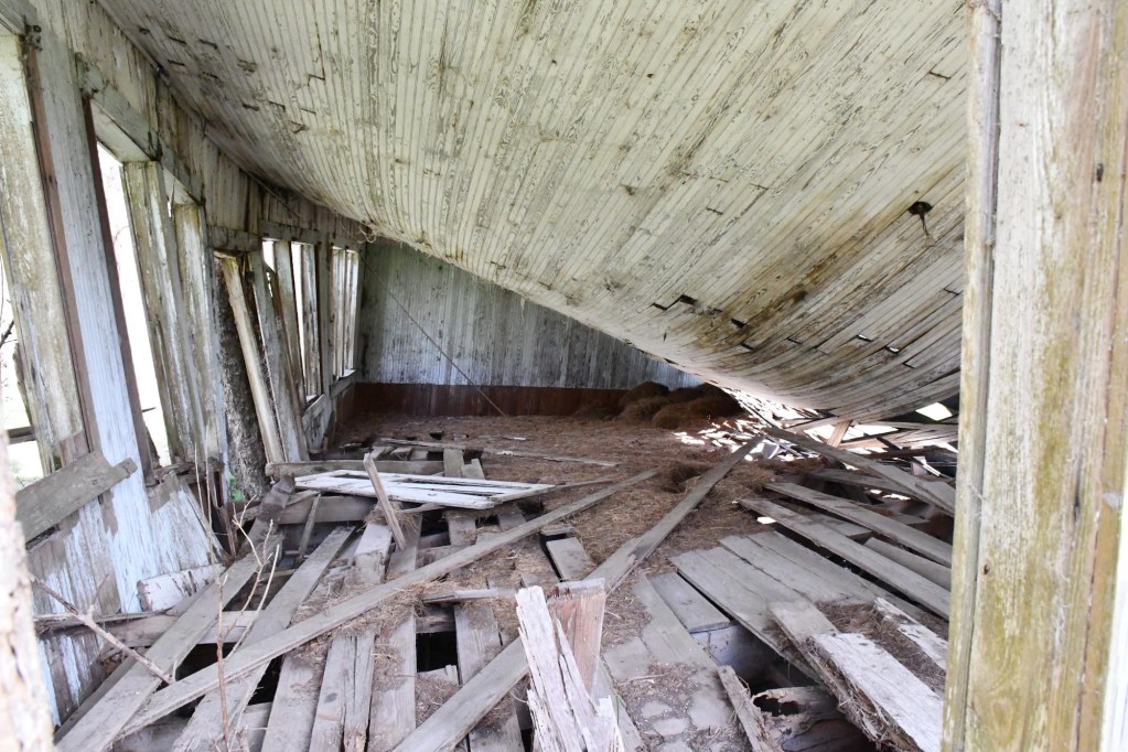 The interior of a deteriorating wooden structure. Floorboards are loose and scattered throughout the room. The ceiling has fallen, angling downward toward the right side of the room.