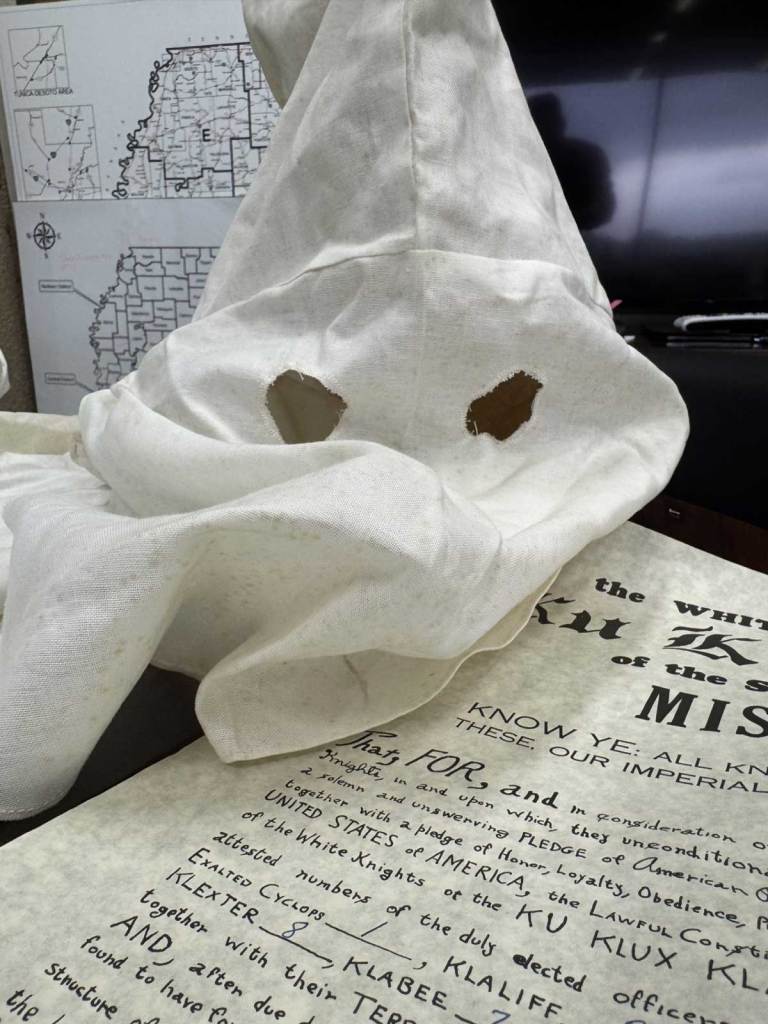 A white hooded mask sits on top of A sealed handwritten document titled 'The White Knights of the Ku Klux Klan of the Sovereign Realm of Mississippi'