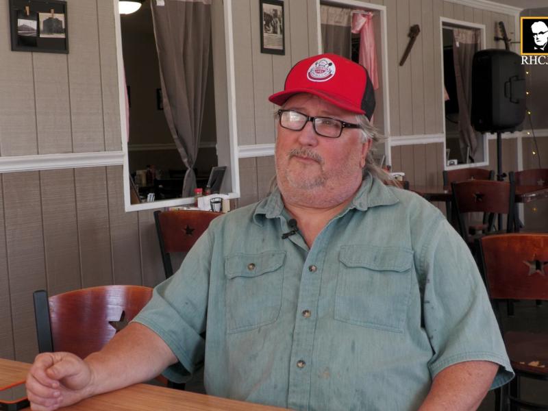 Alan Triplett sits at a table inside Lumberton Cafe wearing a red cap and glasses during an interview