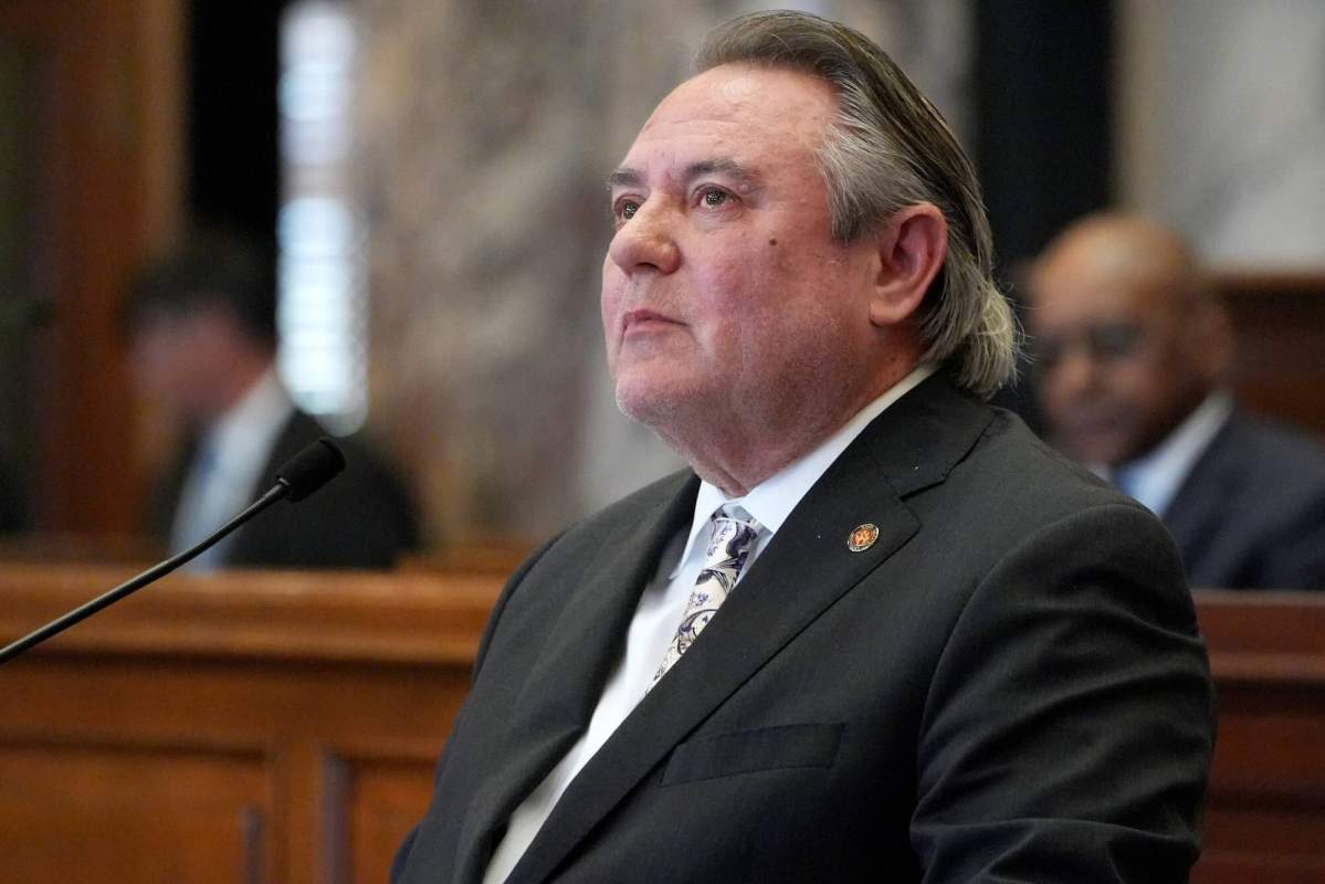 A man in a black suit sits at a mic, looking up