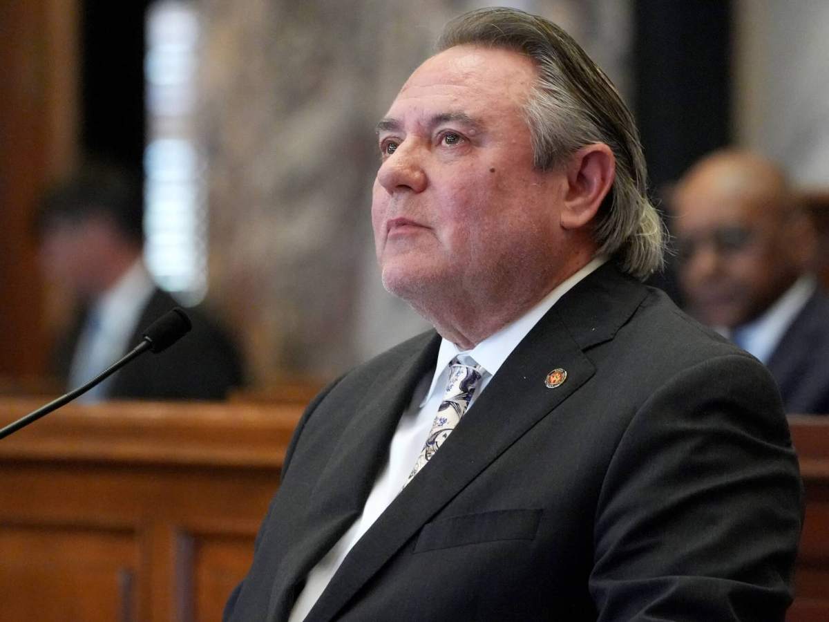 A man in a black suit sits at a mic, looking up