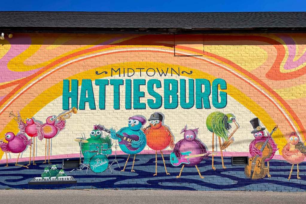 A colorful warm mural on the side of a building that reads 'Midtown Hattiesburg' and depicts birds playing in a band under an orange rainbow