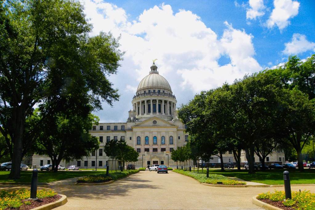 The Mississippi Capitol building in Jackson seen from down the entrance roadway. It's framed by large green trees on either side