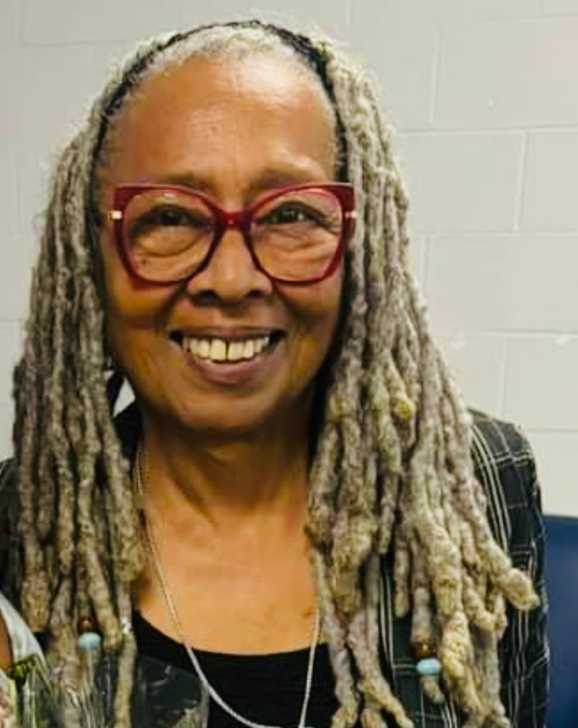 A woman in red glasses and long greying dreadlocks smiles