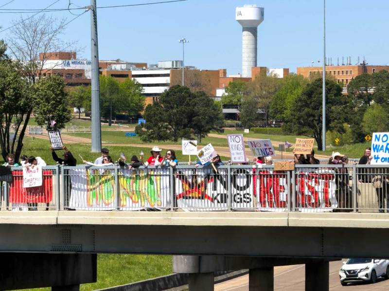 A bridge over the interstate is lined with protestors holding signs that read 'HONK!' 'NO KINGS' 'RESIST'