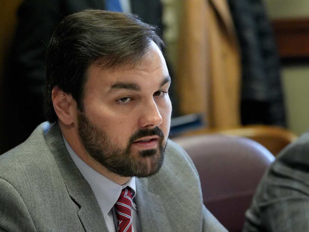 A closeup of Noah Sanders in a light grey suit speaking at a meeting