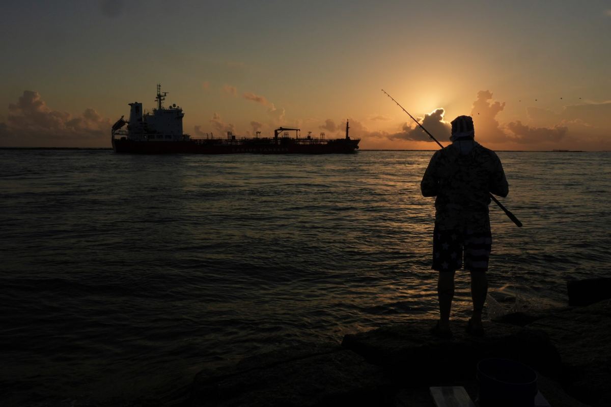 A man fishing with an oil tanker traveling in the sea in the background. The sun is setting.
