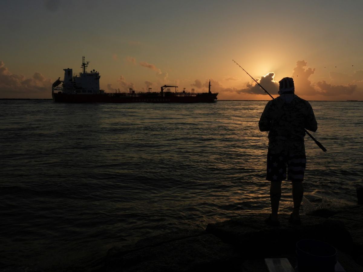A man fishing with an oil tanker traveling in the sea in the background. The sun is setting.