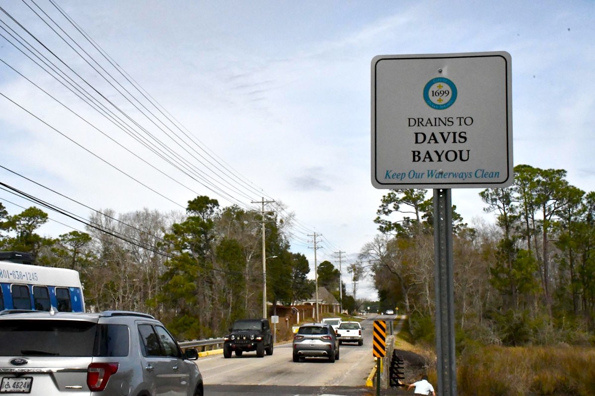 Cars drive across a two-lane bridge near a roadside sign reading “Drains to Davis Bayou: Keep Our Waterways Clean.”