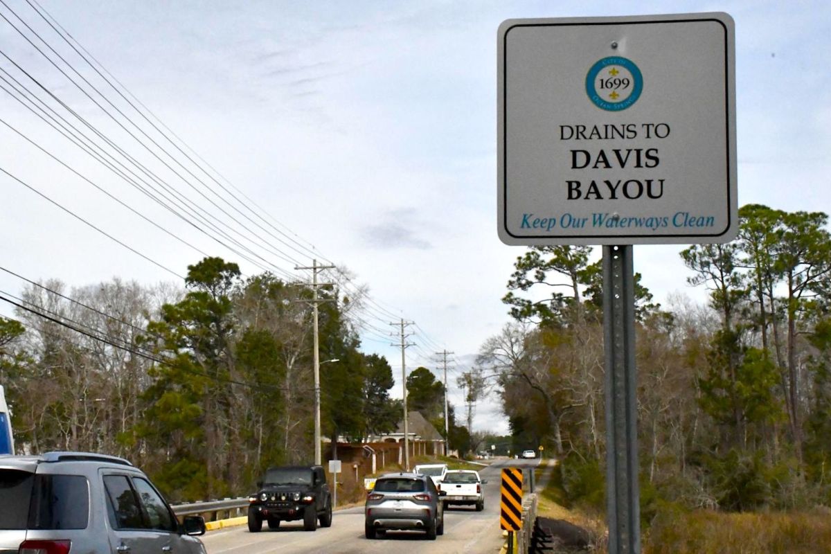 Cars drive across a two-lane bridge near a roadside sign reading “Drains to Davis Bayou: Keep Our Waterways Clean.”
