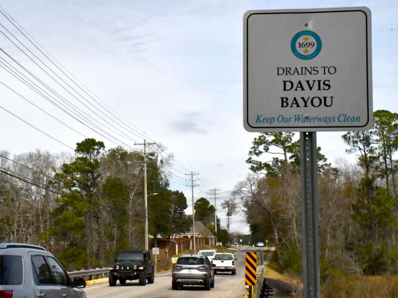 Cars drive across a two-lane bridge near a roadside sign reading “Drains to Davis Bayou: Keep Our Waterways Clean.”