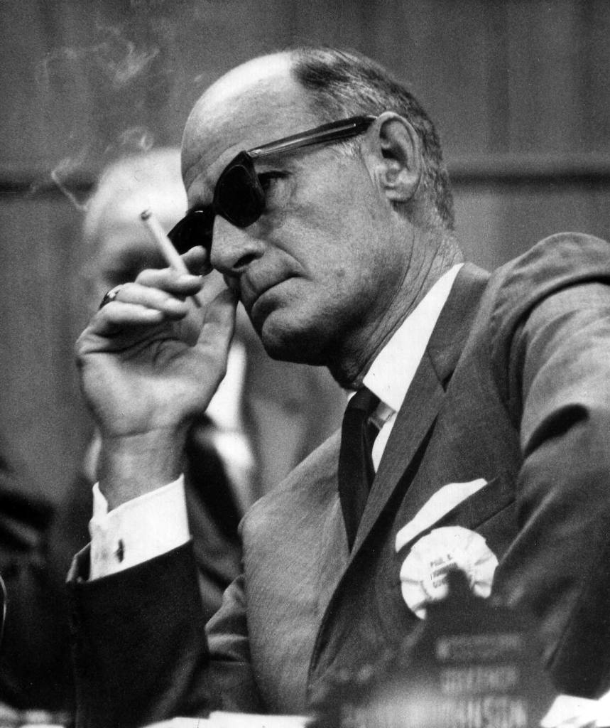 A black and white photo of Paul B. Johnson in a meeting, leaning on one elbow and smoking while listening on