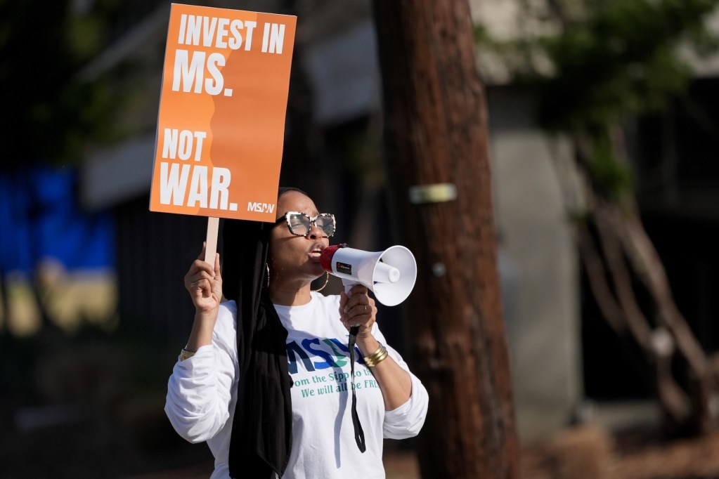A woman speaks through a megaphone while holding up an orange sign that reads 'Invest in MS. Not War.'
