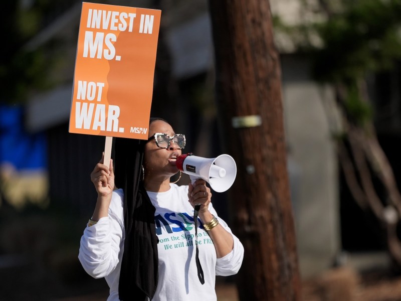 A woman speaks through a megaphone while holding up an orange sign that reads 'Invest in MS. Not War.'