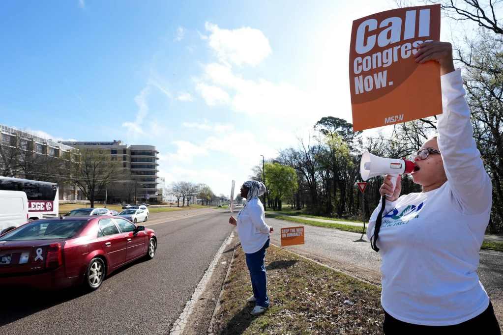 Two people hold signs on the roadside as one woman speaks through a megaphone