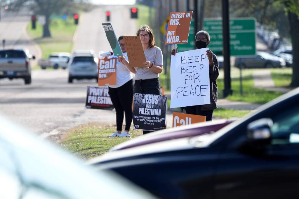 Protestors hold signs by the roadside that read 'Invest in MS' "Beep Beep for Peace'
