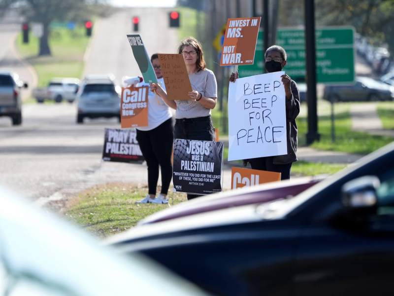 Protestors hold signs by the roadside that read 'Invest in MS' "Beep Beep for Peace'