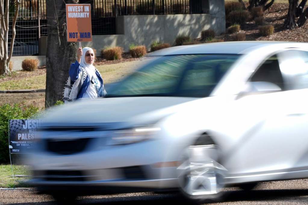 A person holds up a sign by the roadside that reads 'Invest in Humanity'
