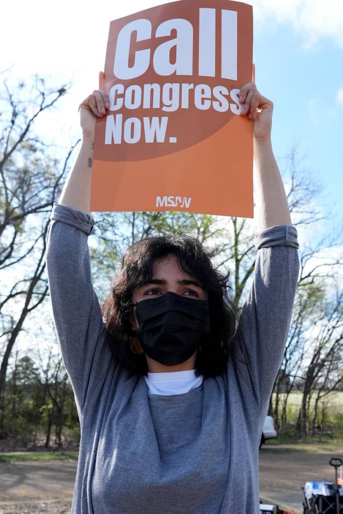 A masked person holds an orange sign that reads 'Call Congress Now'
