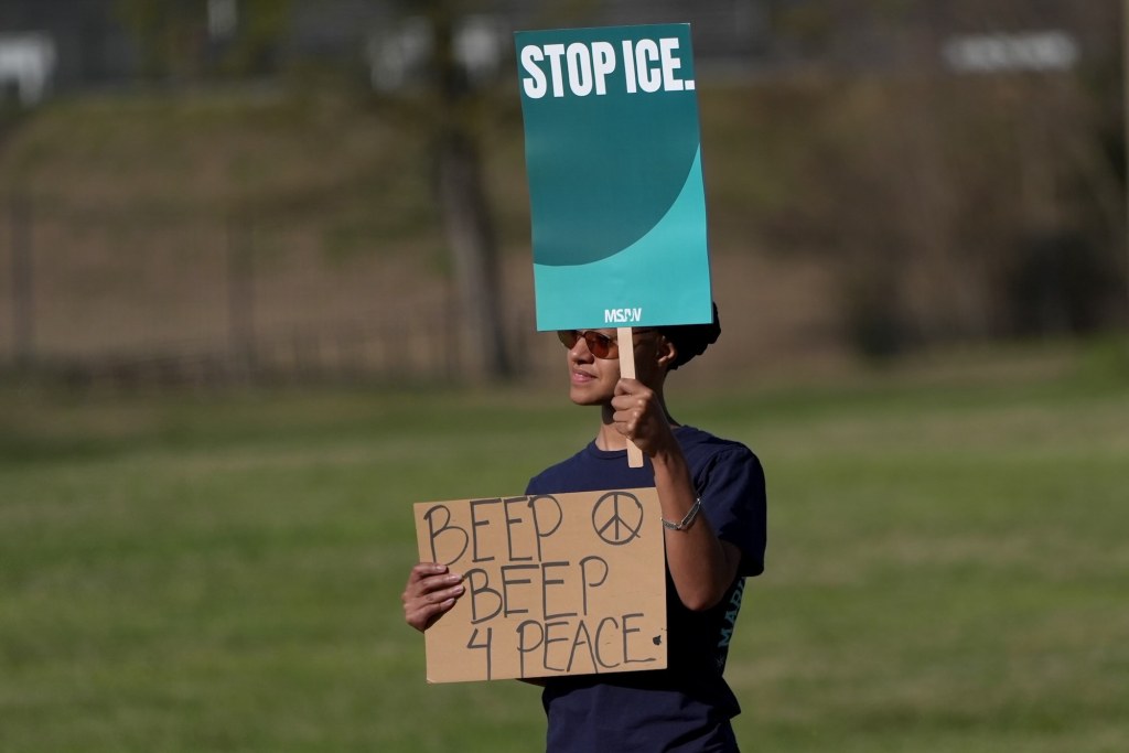 A person holds two signs, one reads 'Stop ICE' and the other 'Beep Beep 4 Peace'