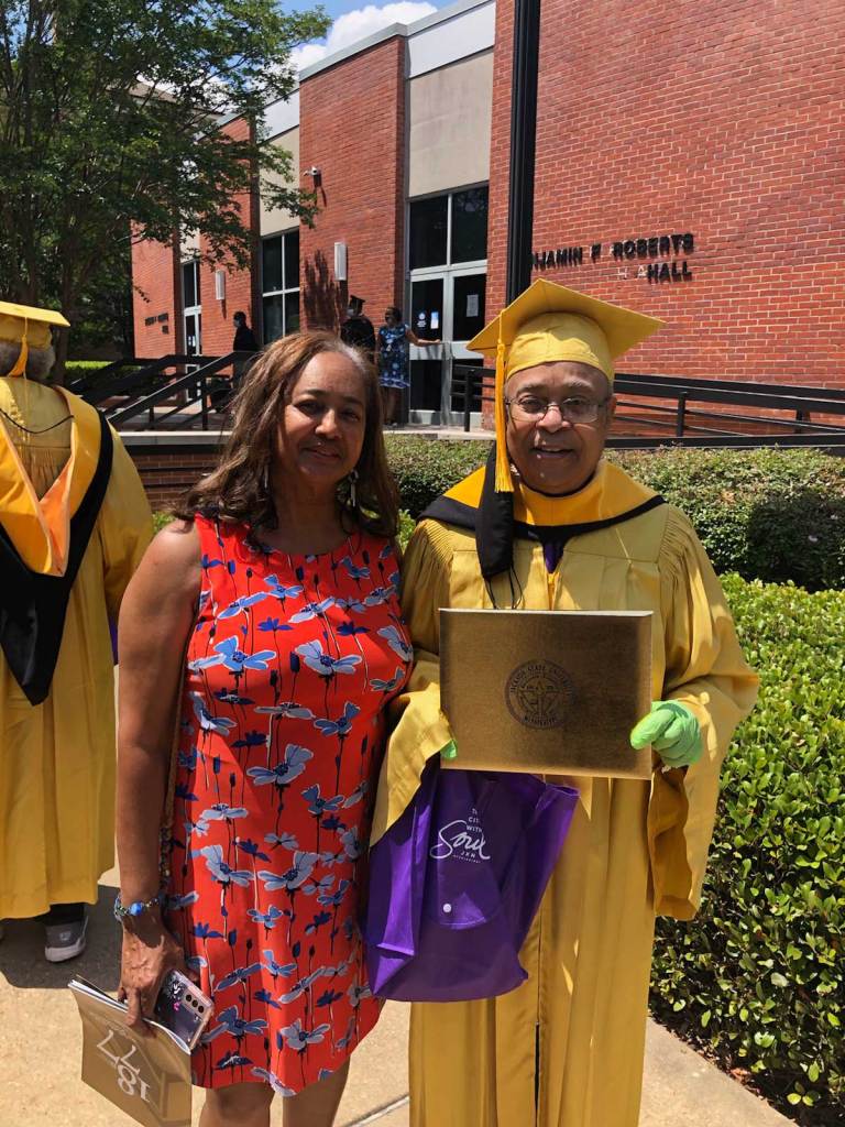 A woman in a red floral dress stands beside an man in yellow cap and gown outside of a campus building