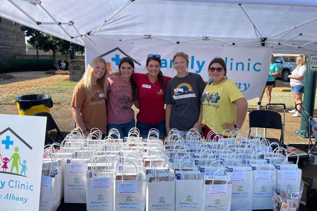 Five people stand under a tent with a sign that reads 'Family Clinic'. A table before them is filled with bags labeled 'Family Clinic of New Albany'