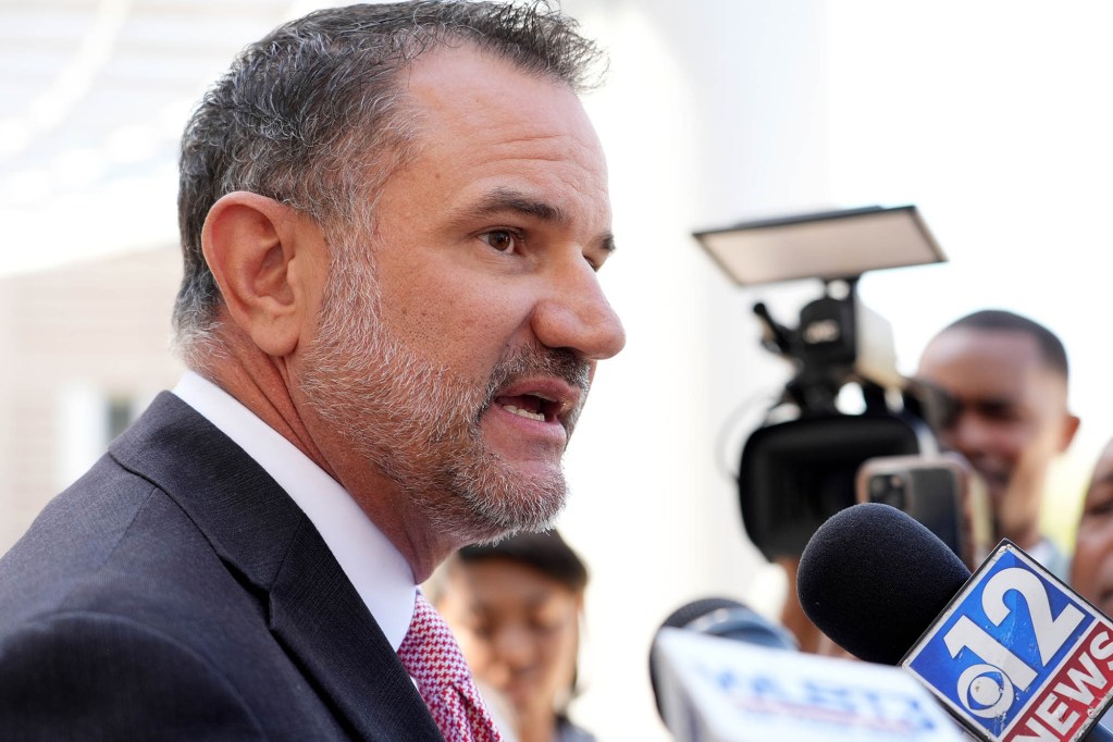 a man in a suit speaks with press members holding cameras
