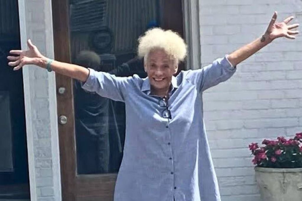 A woman wearing a light blue shirt smiling and holding her arms wide in front of a house