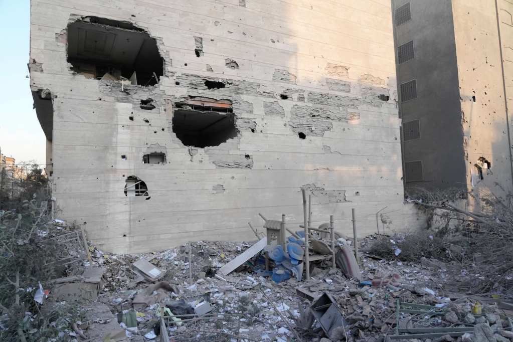 A building’s wall damaged by bombing, with several holes in it. The ground around the building is covered in rubble.