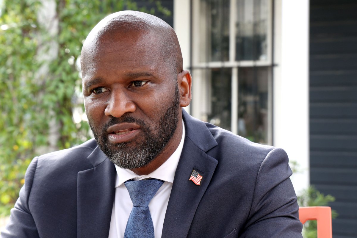 A man in a blue suit with an american flag lapel pin, speaking outside
