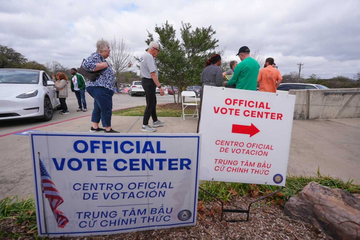 People gather in a line outside at tables. In the foreground are two signs that read “Official Vote Center” with both Spanish and Vietnamese directions.
