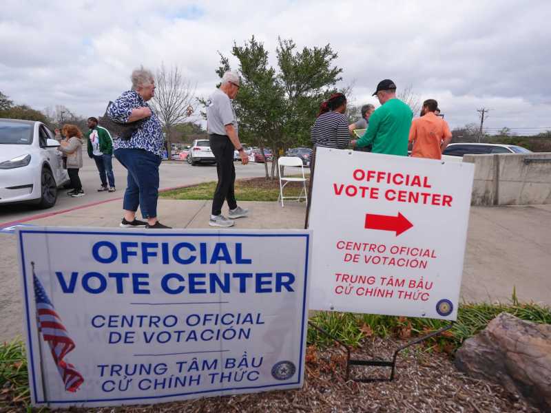 People gather in a line outside at tables. In the foreground are two signs that read “Official Vote Center” with both Spanish and Vietnamese directions.