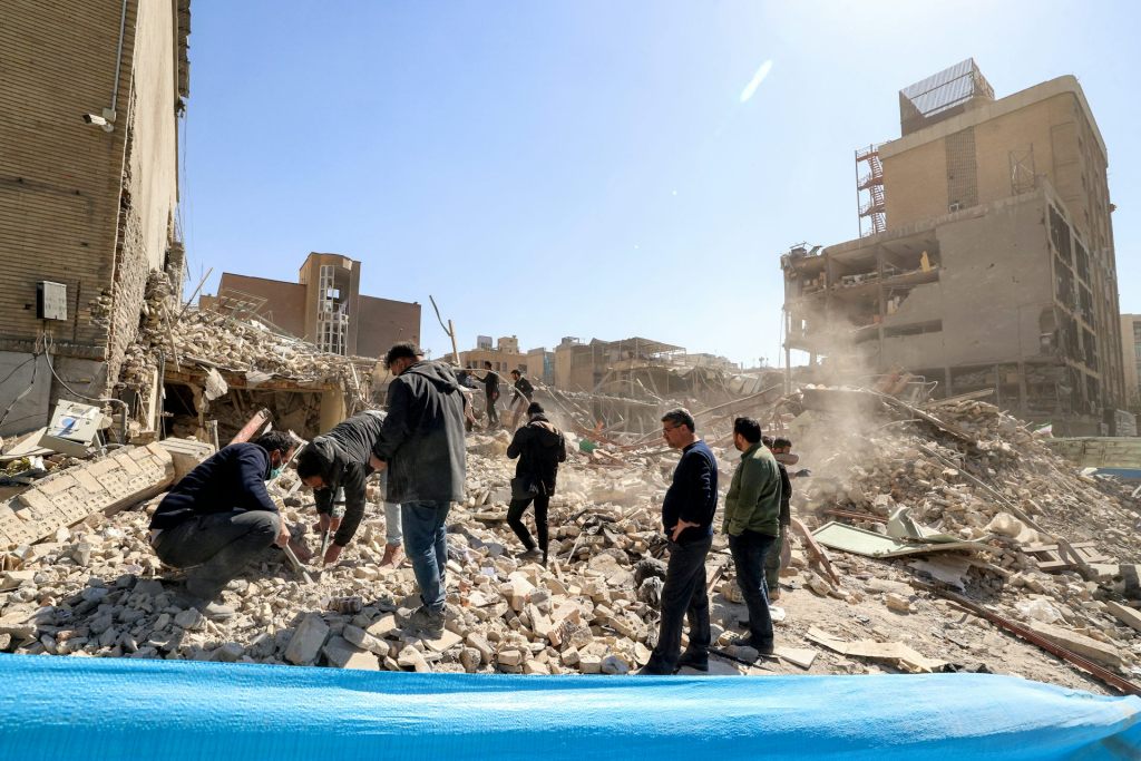 A group of men walk through the remains of a building that collapsed. Several buildings around them are also damaged