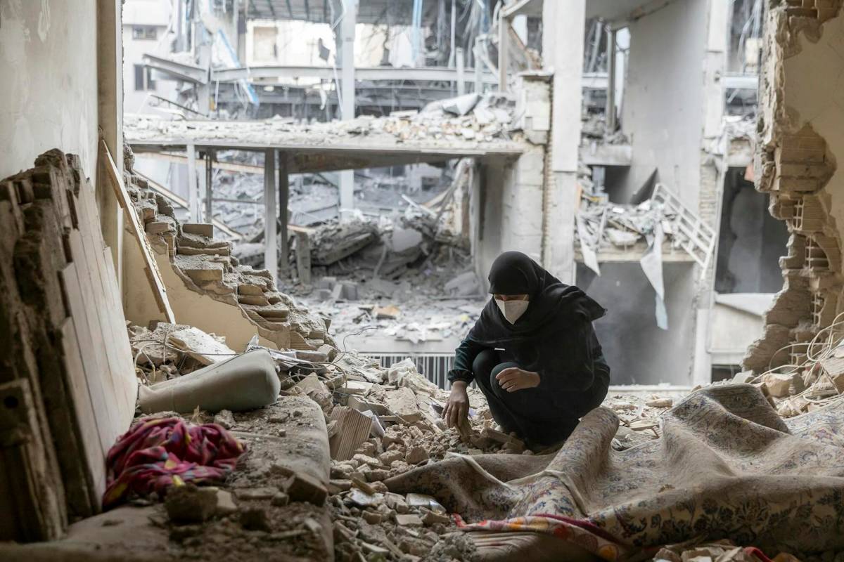A kneeling woman wearing a medical mask sifts through a bombed building