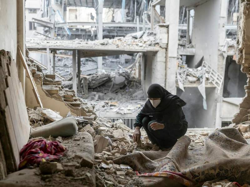 A kneeling woman wearing a medical mask sifts through a bombed building
