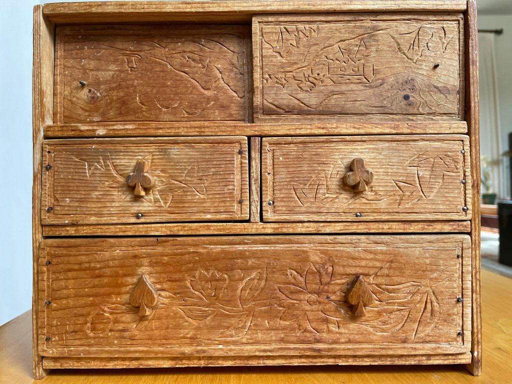A brown wooden chest with handles carved like spades and clubs, with carvings of scenes on the faces of each panel.