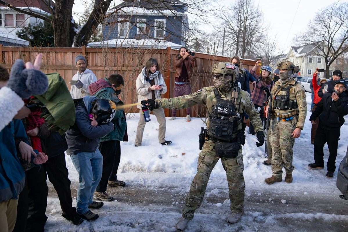 A masked agent sprays pepper spray onto a person in winter clothing. The person is wearing a helmet and looking down to deflect the spray onto the helmet.