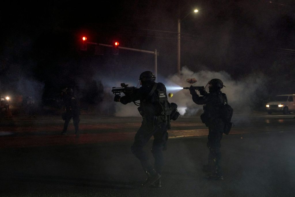 Two police officers dressed in tactical gear aim gun-like weapons into the dark night, with a cloud of tear gas in the background