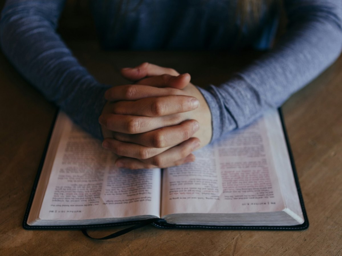A man’s hands clasped and resting on an open book.