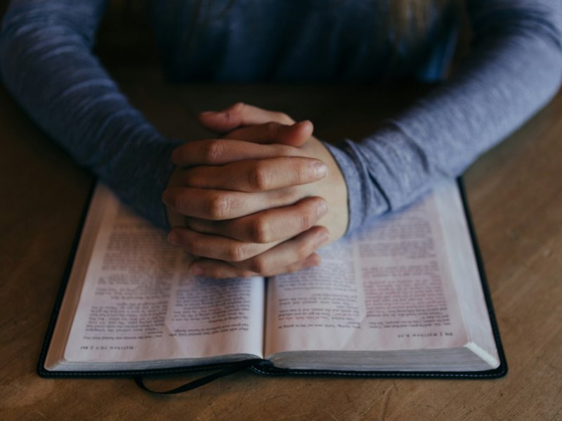 A man’s hands clasped and resting on an open book.
