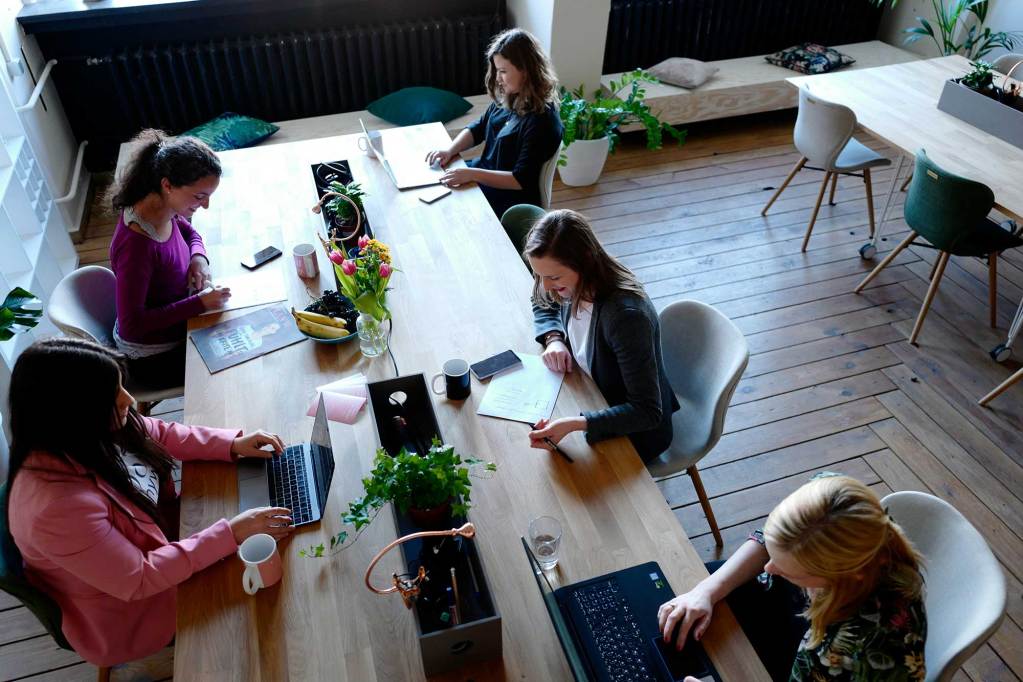 An overhead view of several women working at a long table on either laptops or writing things on paper