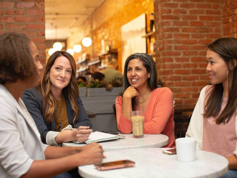 Four women sit around a table in a coffee shop, holding a meeting