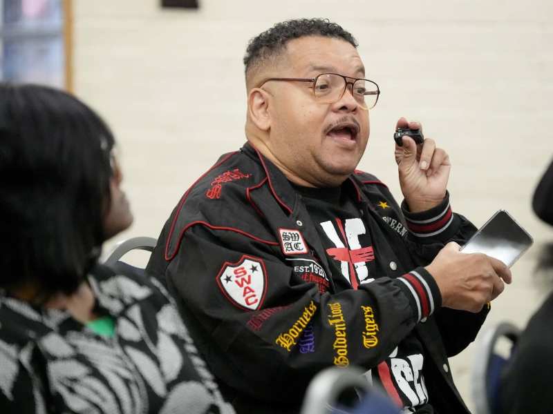 A seated man in a black and red SWAC jacket speaks at a public meeting