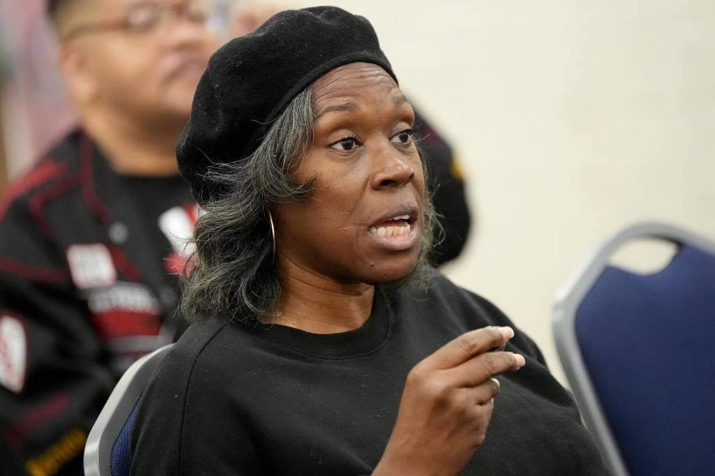 A seated woman in a black beret speaks at a public meeting