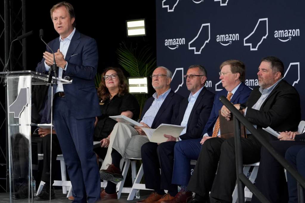 A man in a navy suit speaks at an Amazon press conference, several others seated behind him on stage