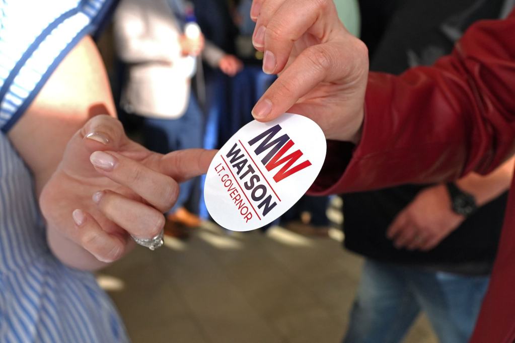 A person in a red jacket hands another person a sticker that reads 'MW Watson Lt. Governor'