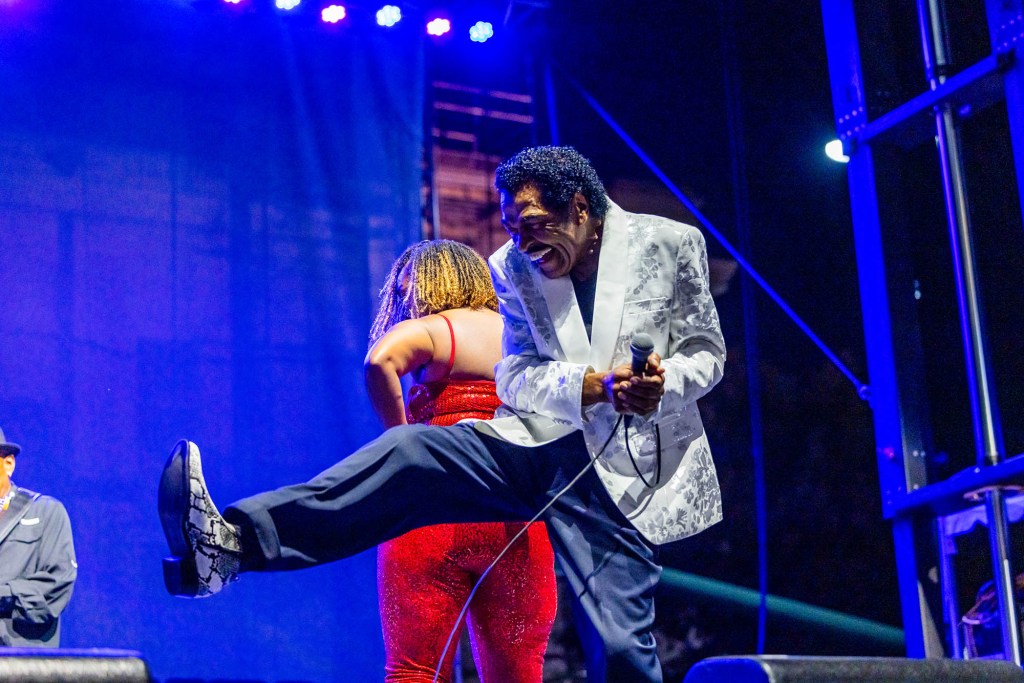 Bobby Rish, in a silver brocade jacket and black pants, dances on a stage lit in blue