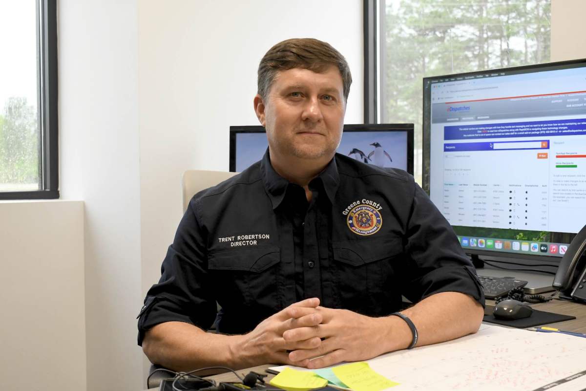 Trent Robertson sits at a desk and smiles at the camera. His shirt reads “Trent Robertson, Director” and displays a Greene County Emergency Management logo. Two computer monitors are behind him.